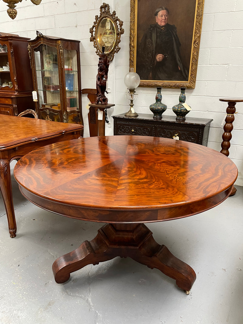 Antique flame mahogany round dining table with a pedestal base and three feet, circumference from the 1840s, seat six persons comfortably. The table is displayed in a room with various other antique furniture pieces in the background.