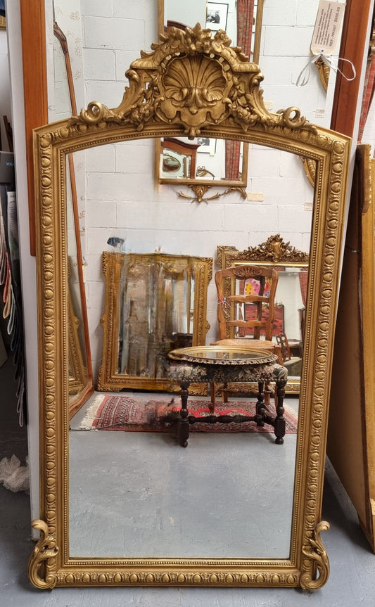 An antique Louis XV style mantle mirror with a decorative top, displayed in a room with various furniture and decorations.