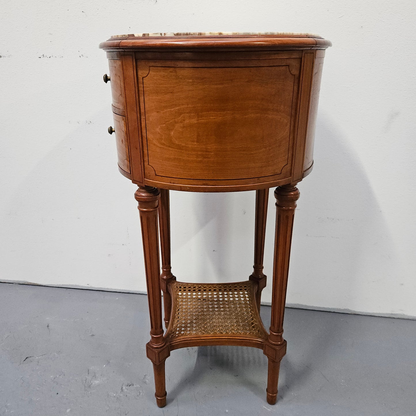 Charming Louis XVI style round side table. Features two drawers, fitted marble top and fluted legs connected by a cane shelf. Sourced directly from France and is in good original detailed condition.