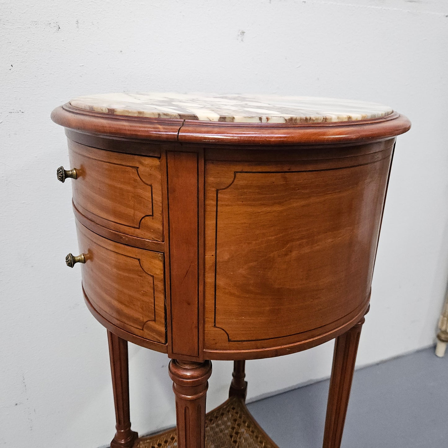 Charming Louis XVI style round side table. Features two drawers, fitted marble top and fluted legs connected by a cane shelf. Sourced directly from France and is in good original detailed condition.