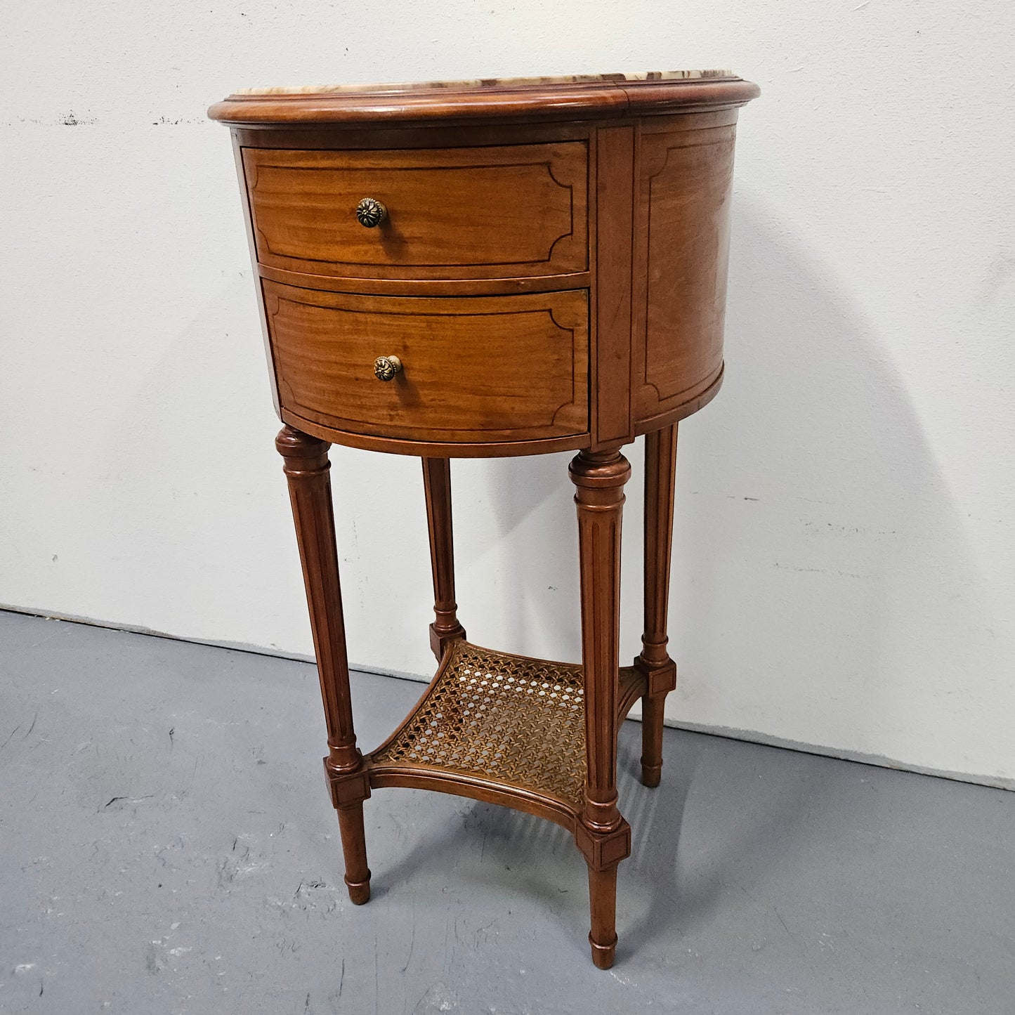 Charming Louis XVI style round side table. Features two drawers, fitted marble top and fluted legs connected by a cane shelf. Sourced directly from France and is in good original detailed condition.