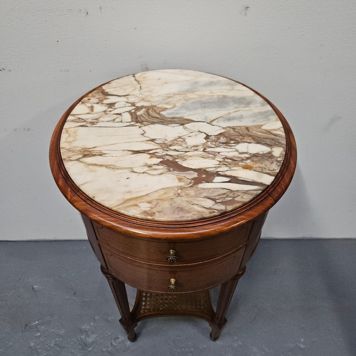 Charming Louis XVI style round side table. Features two drawers, fitted marble top and fluted legs connected by a cane shelf. Sourced directly from France and is in good original detailed condition.