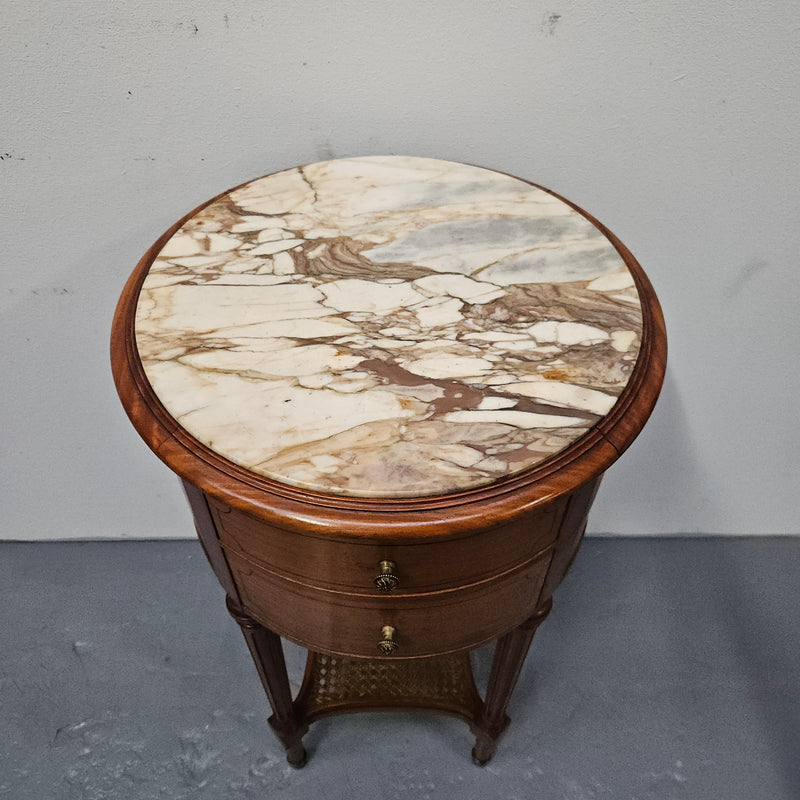 Charming Louis XVI style round side table. Features two drawers, fitted marble top and fluted legs connected by a cane shelf. Sourced directly from France and is in good original detailed condition.