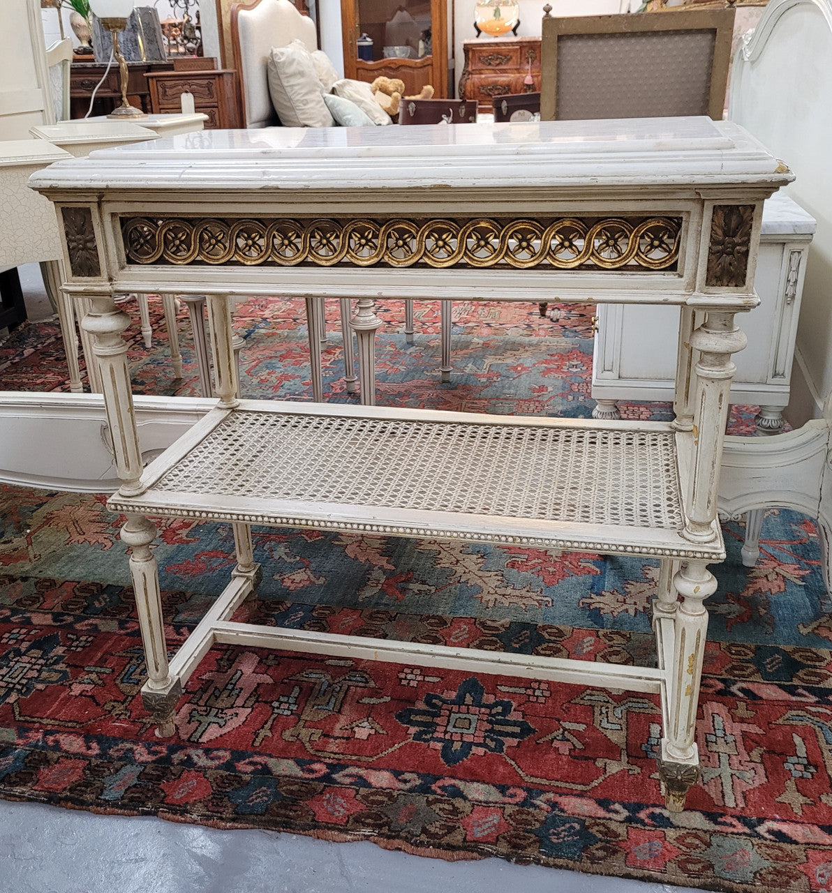 A fabulous painted French Louis XVI style white marble top console table. It has lovely carved detail with gilt highlights and a cane shelf. It has been sourced from France and is in good original condition.
