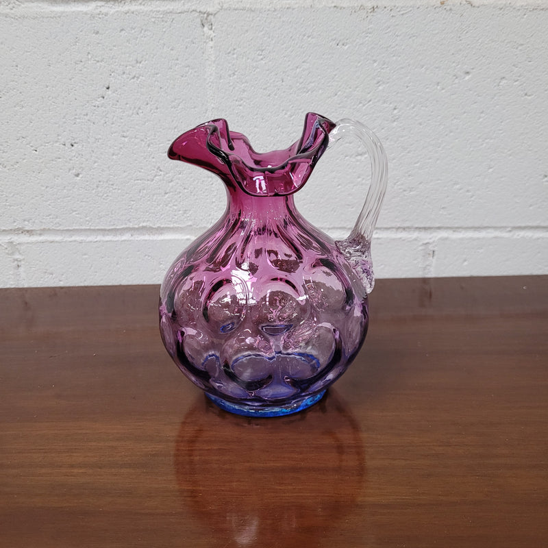 Elegant Fenton Mulberry / Amethyst glass jug in the inverted thumbprint style.  It has a clear stretched glass handle and a ruffled top with a touch of blue at the base. In excellent condition. 