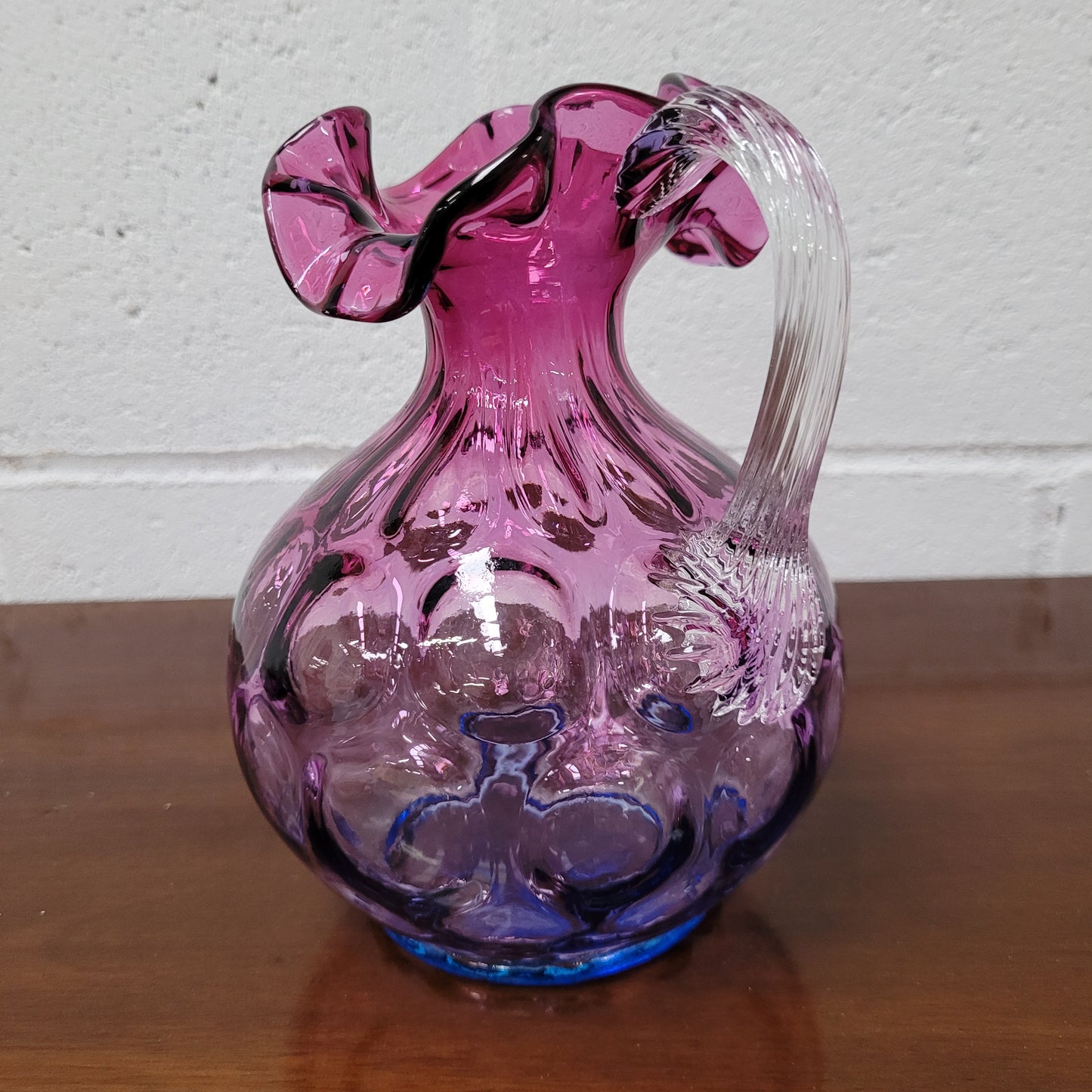 Elegant Fenton Mulberry / Amethyst glass jug in the inverted thumbprint style.  It has a clear stretched glass handle and a ruffled top with a touch of blue at the base. In excellent condition. 