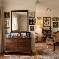 A large, elegant room with warm cream walls, featuring a dark wood dresser under a large mirror, a window with potted plants, and a vintage wooden writing desk on patterned rugs.