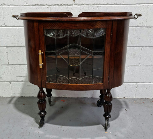 Oak Tudor auto trolley with beautiful leadlight glass. It is on wheels and the door open up to a cabinet with one shelf. In good original detailed condition.